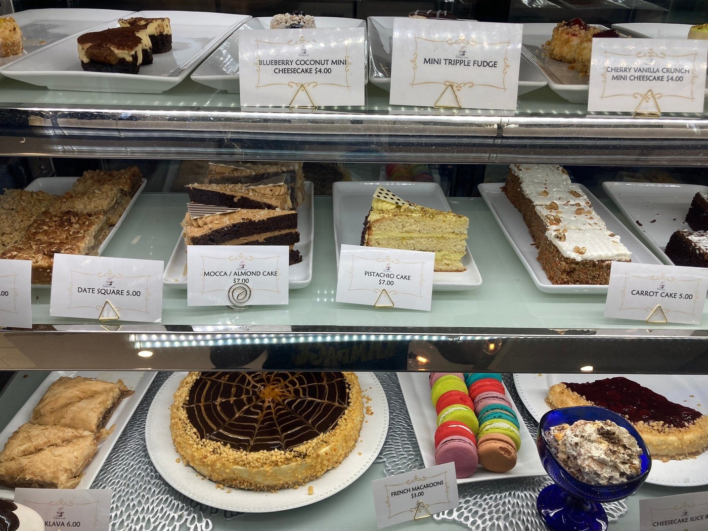 Glass dessert display with individual cakes, layered slices, crumb-topped squares, pastries and colourful macarons
